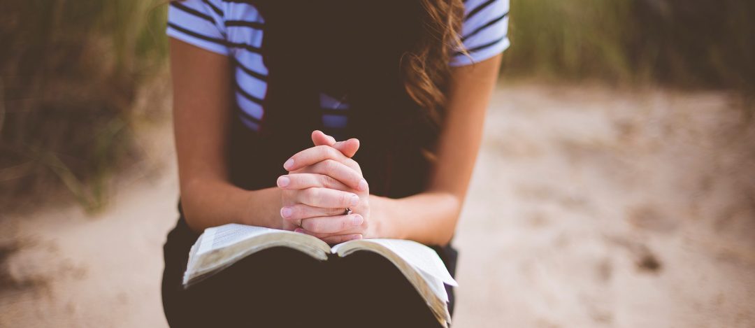 woman in stripey T shirt and dress, sat with the bible on her lap and hands in a position of prayer