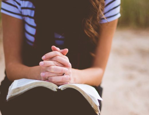 woman in stripey T shirt and dress, sat with the bible on her lap and hands in a position of prayer