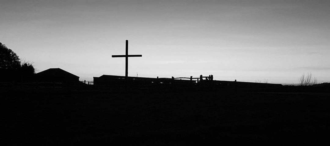 Black and white image of a cross on the hillside with pale sky behind