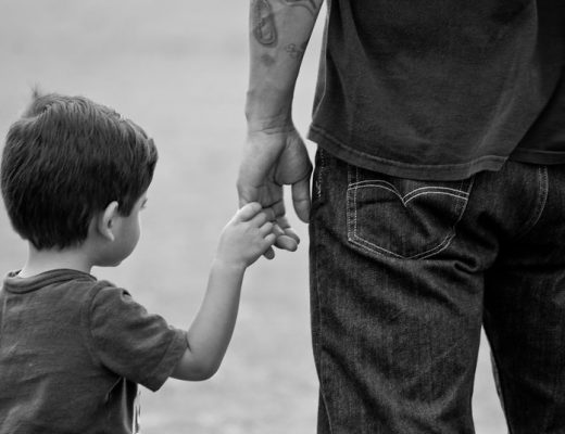 Black and white image of a small child holding his Father's hand, taken from the rear.