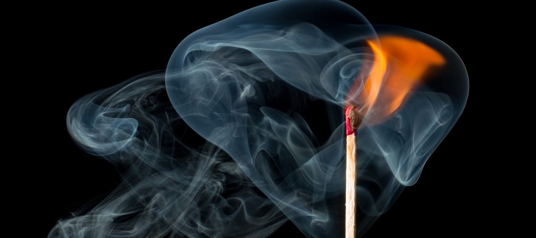 Black background with a match that has been lit, upright with a flame, surrounded by smoke swirling.