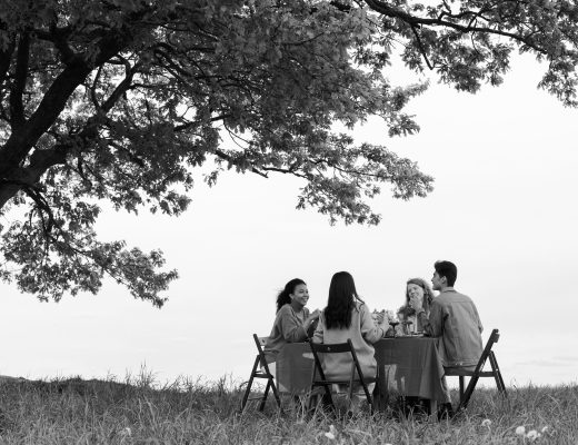 black and white image, of a group of people sat at a table laid for dinner, outside, next to a large tree.