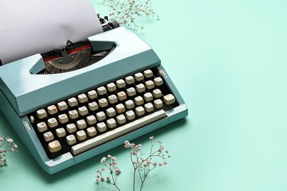 Pale green background, old blue typewriter to the left, with some small white flowers around it.